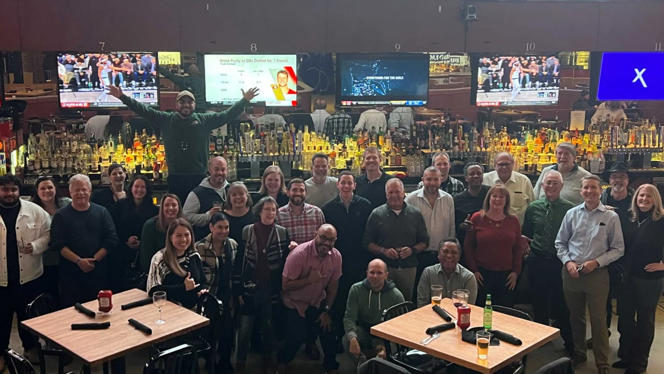 A group of people posing for a photo in a bar with multiple TV screens in the background. One person stands on the bar with arms raised. Tables with drinks are visible in the foreground.