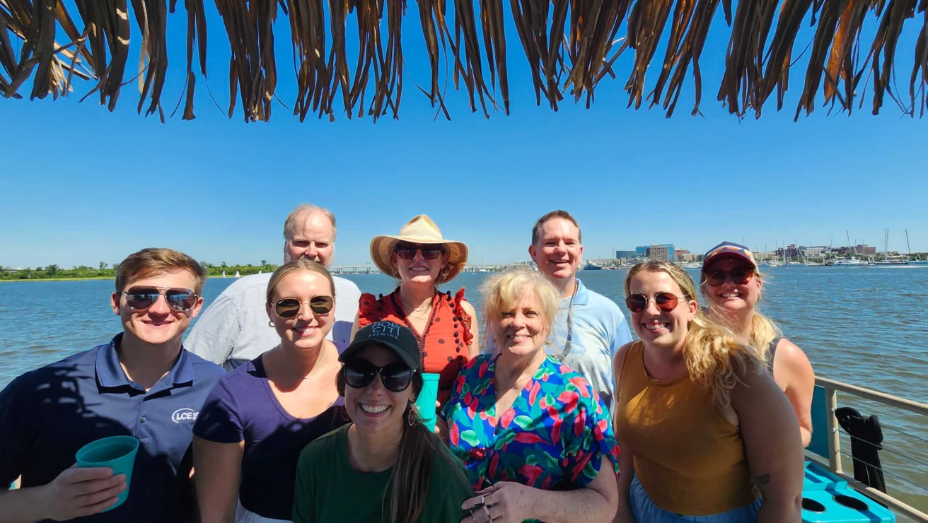 A group of people smiling and standing together under a thatched roof by the water on a sunny day.