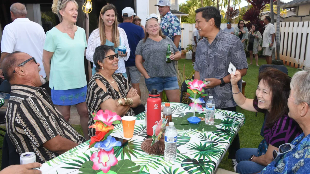 People enjoying a casual outdoor gathering, sitting and standing around a table with tropical decorations.
