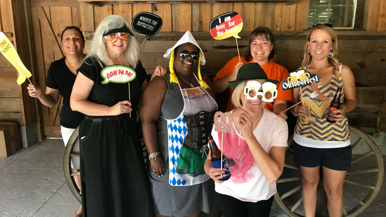 Six people standing indoors, holding playful signs and props, pose for a group photo. They are in a rustic setting with a wooden backdrop.