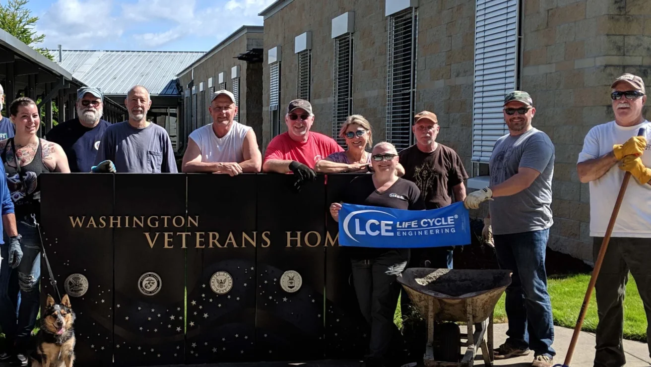 Group of people standing outside, some holding a banner reading "LCE Life Cycle Engineering," in front of a "Washington Veterans Home" sign. A dog sits beside them, and tools are visible.