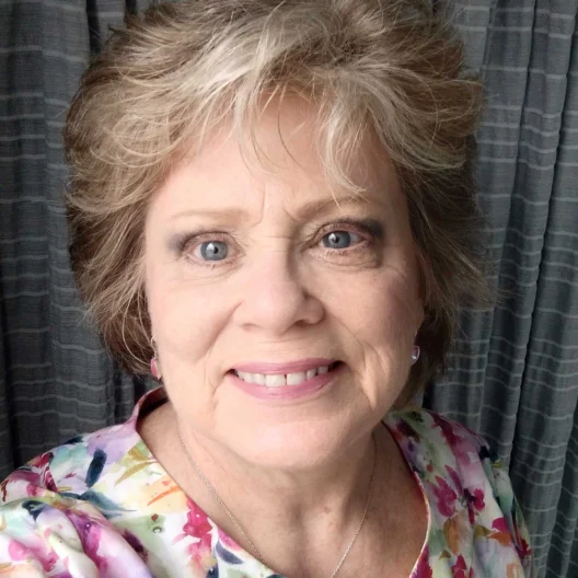 A woman with short, layered hair smiles at the camera, wearing a floral top, standing in front of gray curtains.
