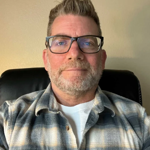 Man with glasses and a short beard, wearing a plaid shirt and a white t-shirt, seated against a beige wall.