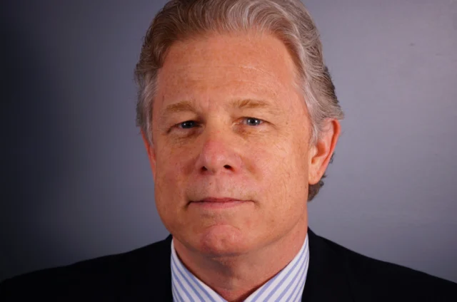 A man with gray hair wearing a suit and tie, standing against a plain gray background.