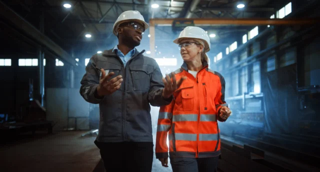 A group of six men standing in a factory, engaged in conversation. One man in a white shirt holds a tablet. They are surrounded by industrial machinery and equipment.