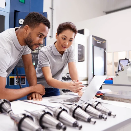 Two engineers in a factory setting examine a laptop, surrounded by metal components and machinery.