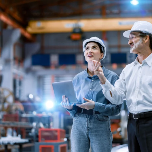 Two engineers in hard hats discuss in a factory, one holding a laptop.