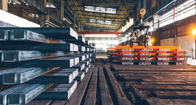 Stacks of metal beams in an industrial warehouse, with some beams heated and glowing orange, under overhead cranes and high ceilings.