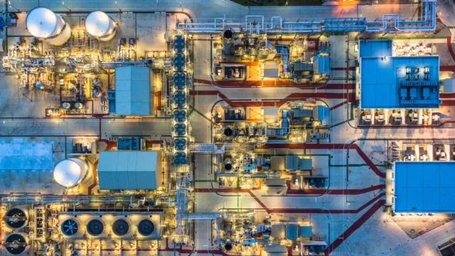 Aerial view of an industrial facility with numerous tanks, pipes, and buildings, illuminated at night, showing an organized layout with connecting walkways and pathways.