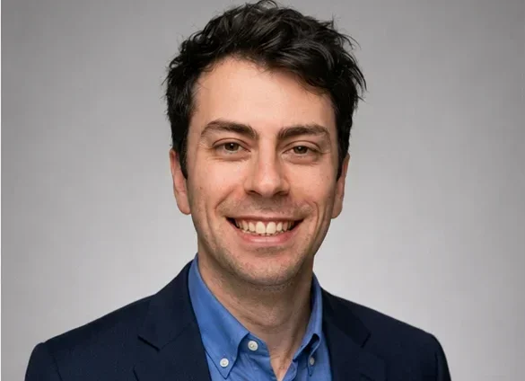 A man with short dark hair smiles at the camera, wearing a blue dress shirt and a dark blazer. The background is plain and light gray.