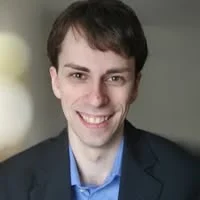 A man with short brown hair wearing a dark blazer and light blue shirt smiles at the camera against a blurred indoor background.