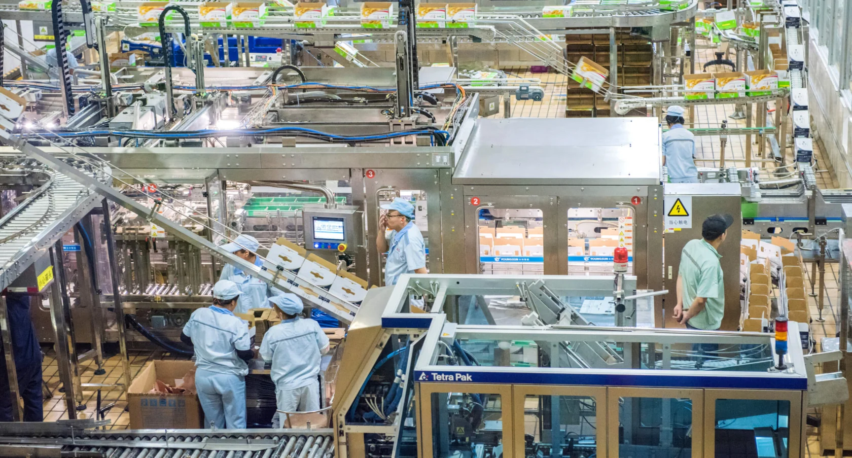 Workers in white uniforms operate machinery on a factory floor, surrounded by conveyor belts and packaged goods, in a busy production environment.