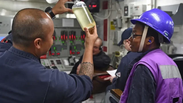 Two workers in a control room, one holding a bottle with yellow liquid. They wear protective gear, including a hard hat and earplugs. Control panel with lights in the background.