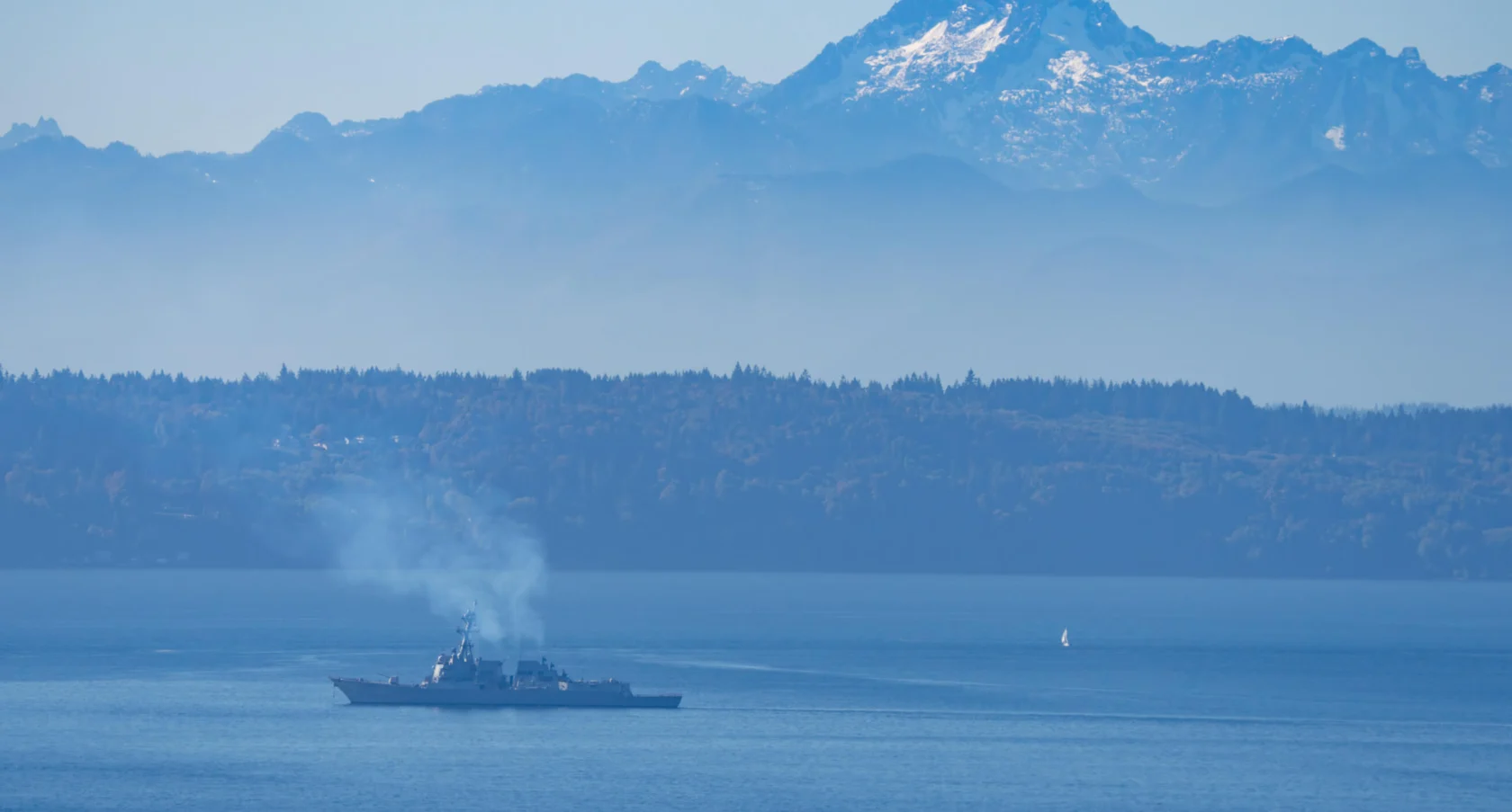 A naval ship sails across a calm blue sea with smoke rising from it. In the background, there are misty mountains under a clear blue sky.