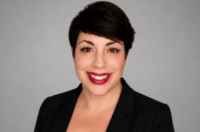 Woman with short dark hair, wearing a black blazer and top, smiling against a plain grey background.