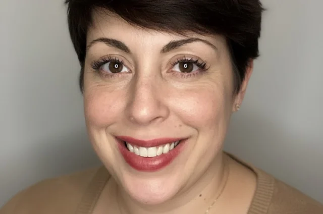 A woman with short dark hair and a beige sweater smiles at the camera against a plain light background.