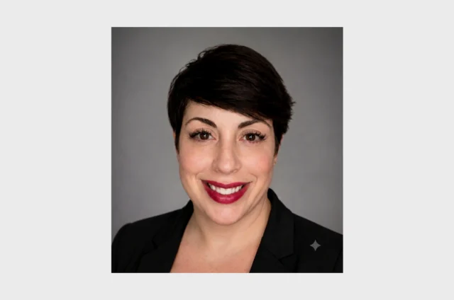 A person with short dark hair and a black blazer smiles at the camera against a plain gray background.