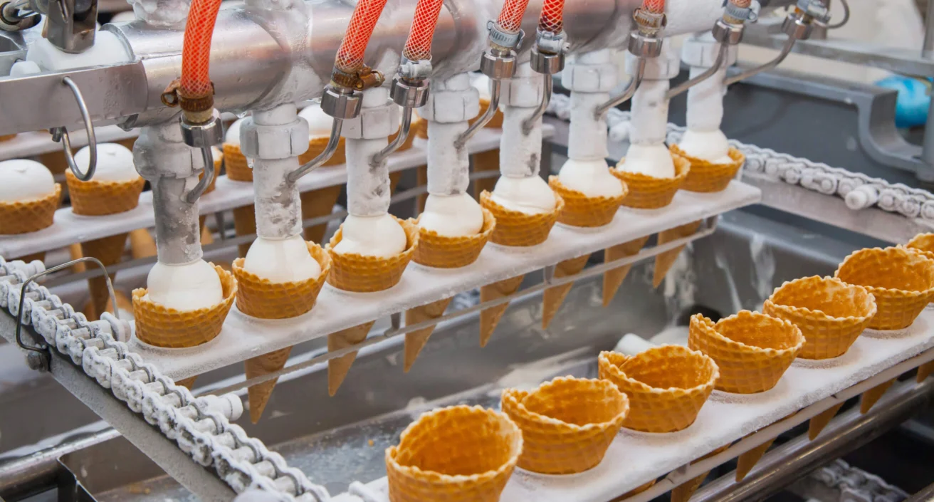 A row of ice cream cones being filled with soft serve by a machine on a production line in a factory setting.