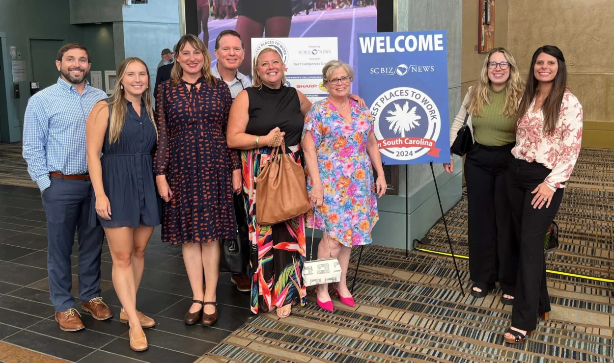 A group of nine people standing together indoors near a sign that reads "Best Places to Work South Carolina 2024.