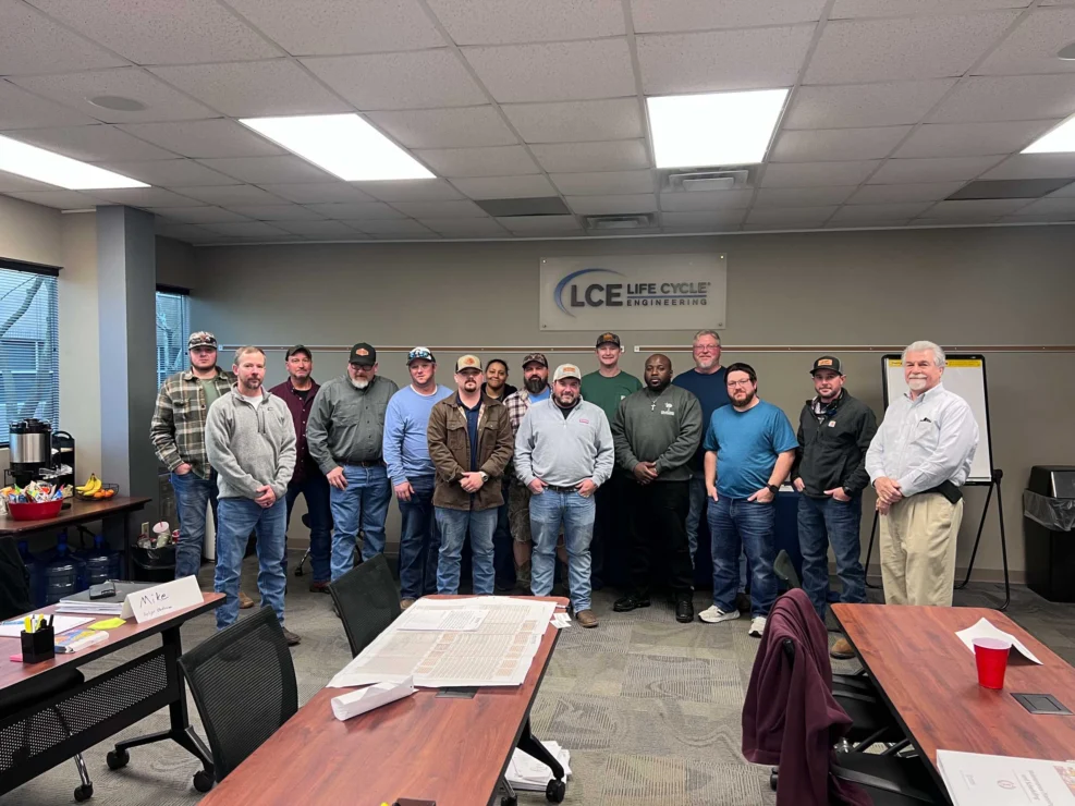 A group of men stand together in a conference room beneath a sign that reads "LCE Life Cycle Engineering." Tables and chairs are visible, along with a whiteboard and office supplies.