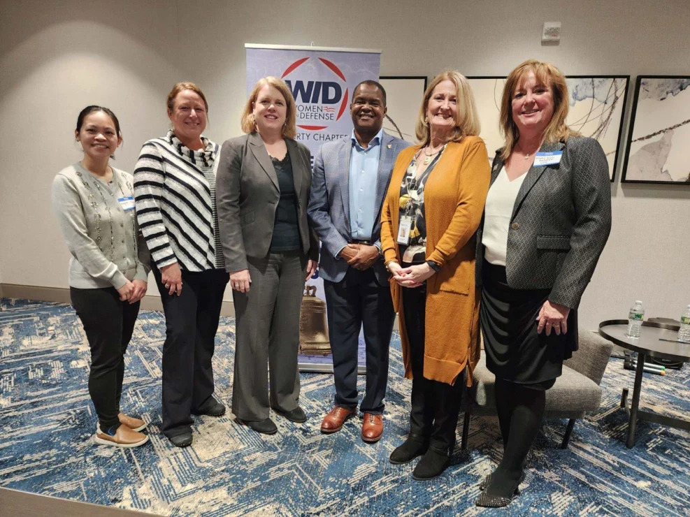 Six people standing in a conference room in front of a "Women in Defense" banner, smiling at the camera.
