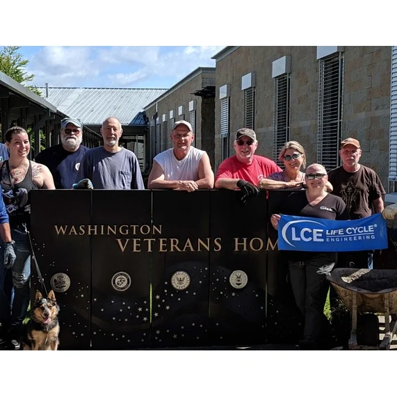 Group of people standing with a "Washington Veterans Home" sign. One holds an "LCE Life Cycle Engineering" banner. A dog is in the foreground. Building and trees are in the background.