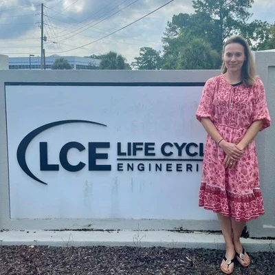 A person in a pink dress stands beside a sign reading "LCE Life Cycle Engineering" with trees and a cloudy sky in the background.