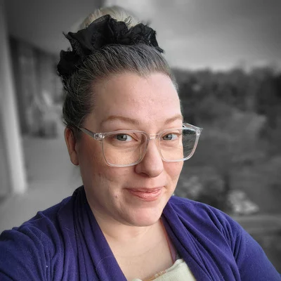 A person with glasses and a bun, wearing a purple top, smiles slightly in a partially desaturated background.