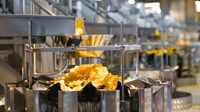 A stainless steel machine dispenses potato chips into a large metal container on an assembly line in a factory setting.