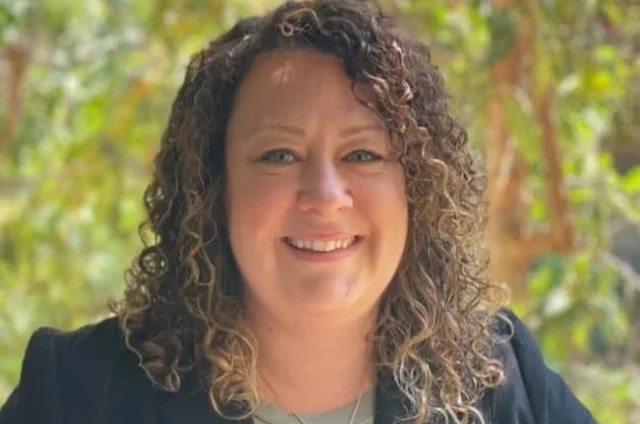 Person with curly hair smiling, wearing a dark blazer and necklace, with greenery in the background.