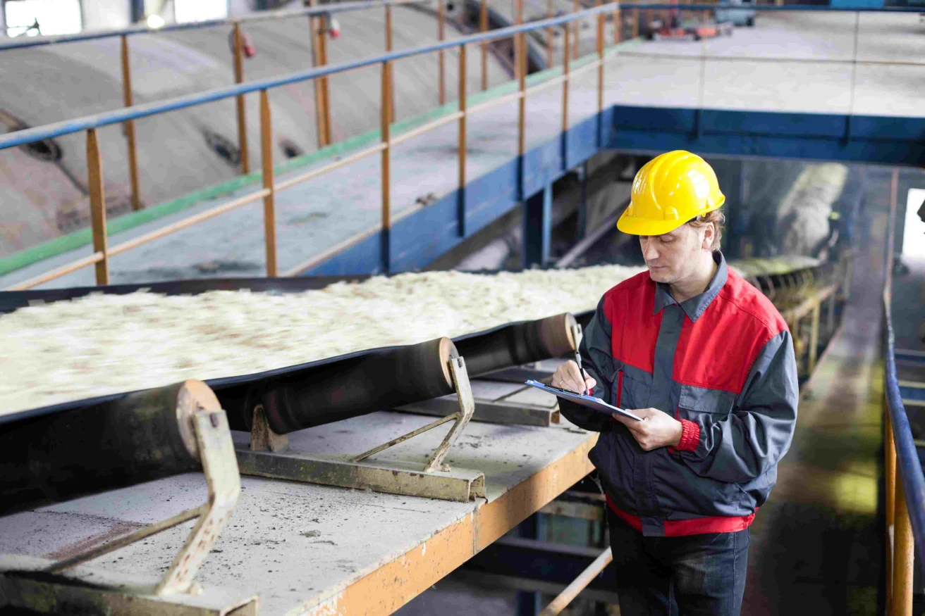 Worker in a hard hat inspects a conveyor belt carrying raw material in an industrial facility, making notes on a clipboard.