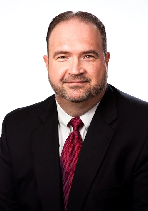 A man with short hair and a beard wearing a dark suit, white shirt, and red tie poses in front of a plain white background.