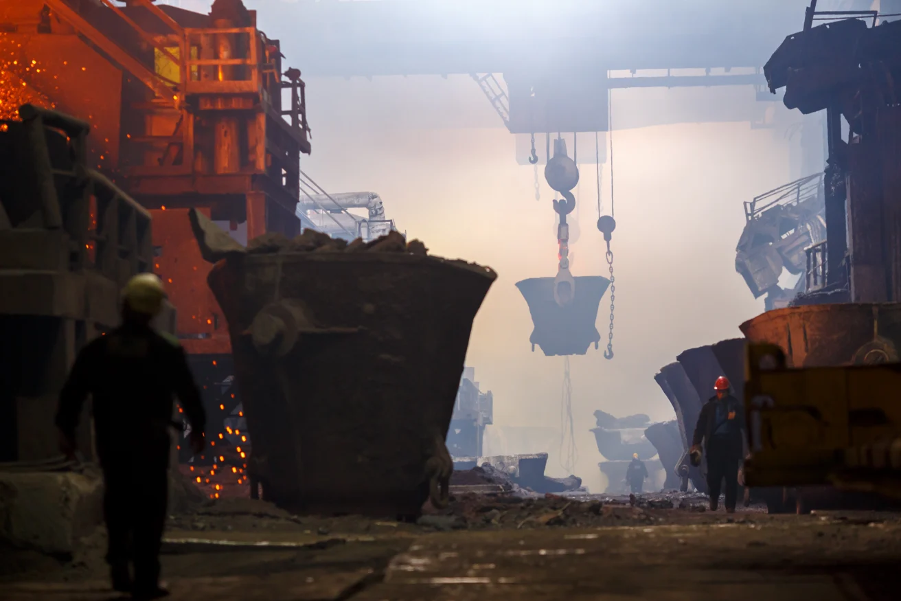 Large industrial foundry interior with workers, molten metal, large crucibles, and overhead cranes amid smoky, illuminated atmosphere.