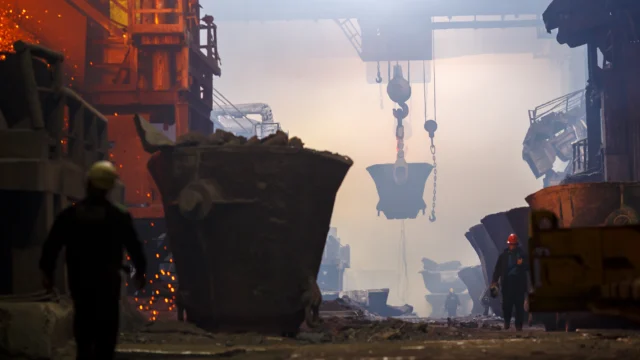 Large industrial foundry interior with workers, molten metal, large crucibles, and overhead cranes amid smoky, illuminated atmosphere.